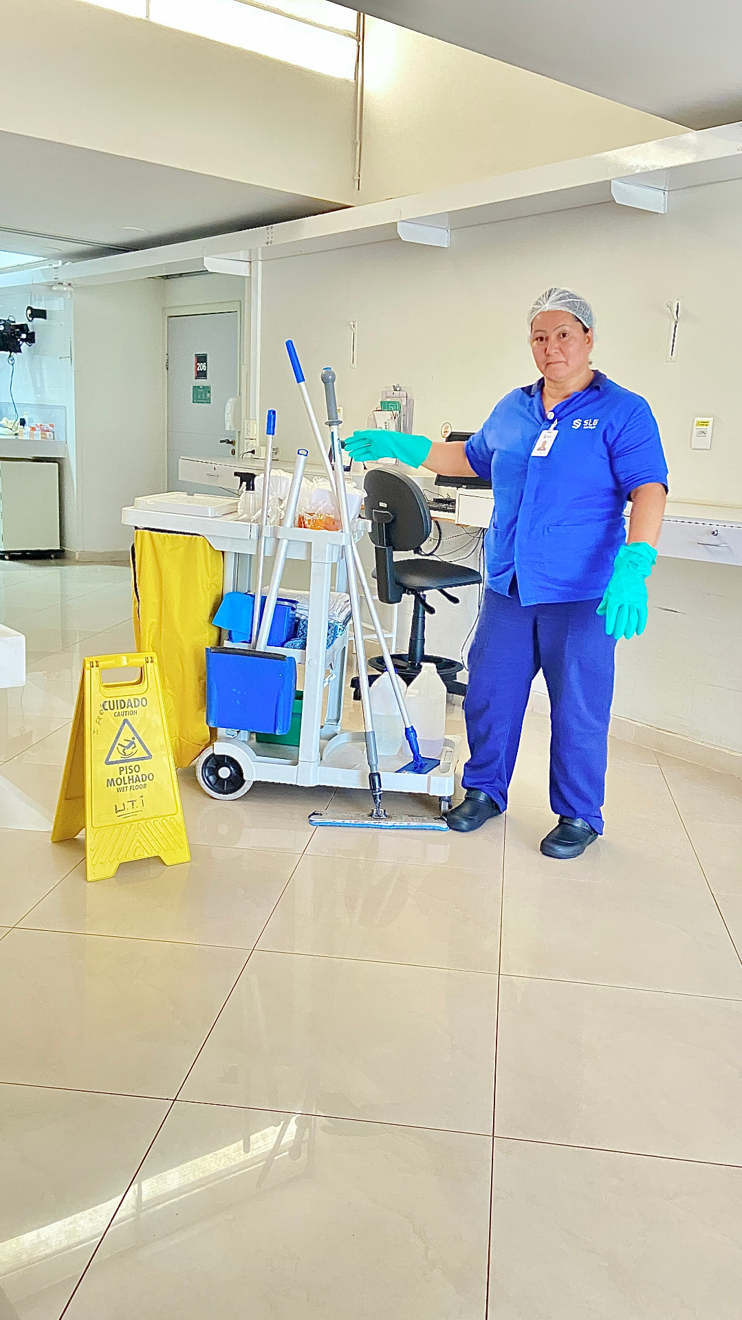 Profissional de limpeza SLG em ambiente hospitalar com carro funcional e sinalização de piso molhado